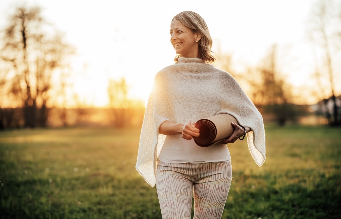 Lächelnd auf einer Wiese im Abendlicht, die Yogamatte unter dem Arm