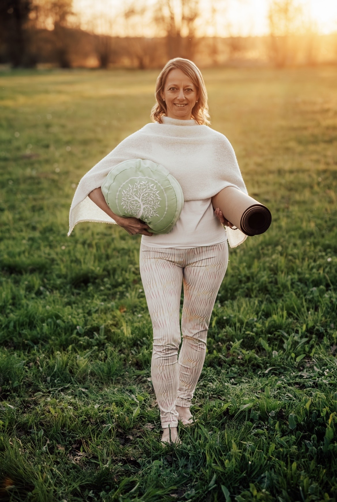 Barfuß im Gras, mit Sitzkissen und Yogamatte im Arm